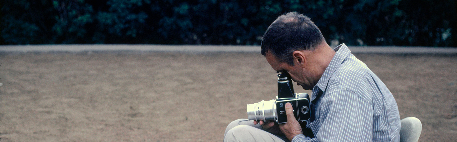 Charles Eames sits on one of his chairs and takes a photo with a Hasselblad camera